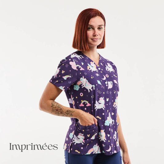 Haut de scrub à thème licorne violette avec manches courtes et encolure en V, orné d'illustrations fantaisistes pastel de licornes, arcs-en-ciel, étoiles, lunes et beignets sur un tissu sombre.